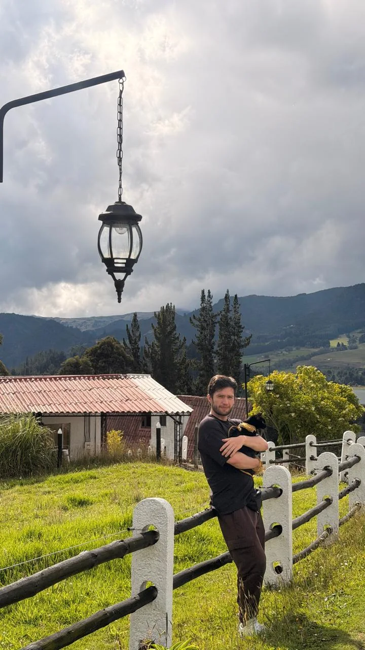 Sergio con un gato en brazos junto a una cerca, rodeado de montañas y vegetación